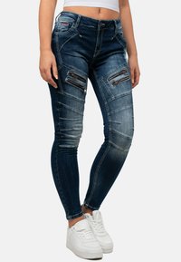 Jeans skinny in denim blu scuro con zone sbiadite, tasche con zip su cosce e dettagli di cucitura angolari. Indossati con sneakers bianche.