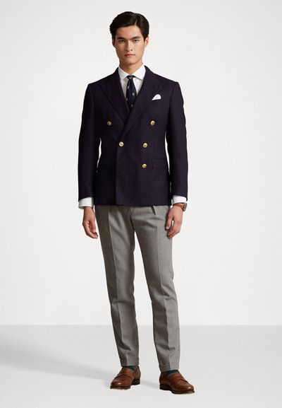 Blazer croisé marine à double boutonnage avec des boutons dorés, chemise blanche, cravate à motifs, pantalon gris sur mesure et chaussures en cuir marron.