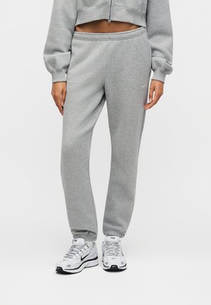 Person mit grauer Jogginghose mit elastischen Bündchen und weißen Nike-Sneakers, stehend vor einem einfachen hellen Hintergrund.