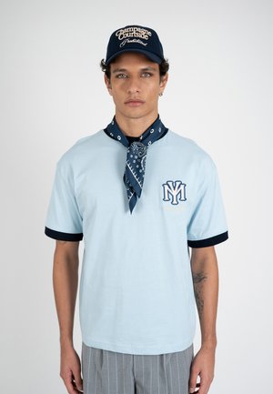 Mann trägt hellblaues T-Shirt mit dunklem Rand, marineblaue Bandana um den Hals, graue Nadelstreifenhose und marineblaue Kappe mit weißem Text.