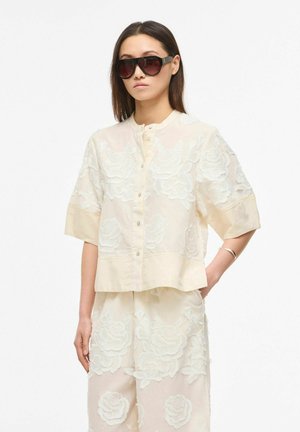 Kvinde iført cremegul blomstret broderet knappet op-top og matchende shorts, med store mørke solbriller og et sølvarmbånd.