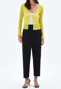 Kobieta w żółtym prążkowanym kardiganie związywanym z przodu, białym topie na ramiączkach, czarnych spodniach z wysokim stanem i czarnych sandałach na obcasie stoi na białym tle.