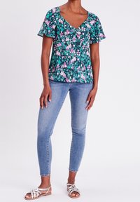 Zelený top s květinovým vzorem, krátkými volánkovými rukávy, výstřihem do V a nařasením v oblasti prsou, zkombinovaný se světlomodrými skinny džínami a sandály.