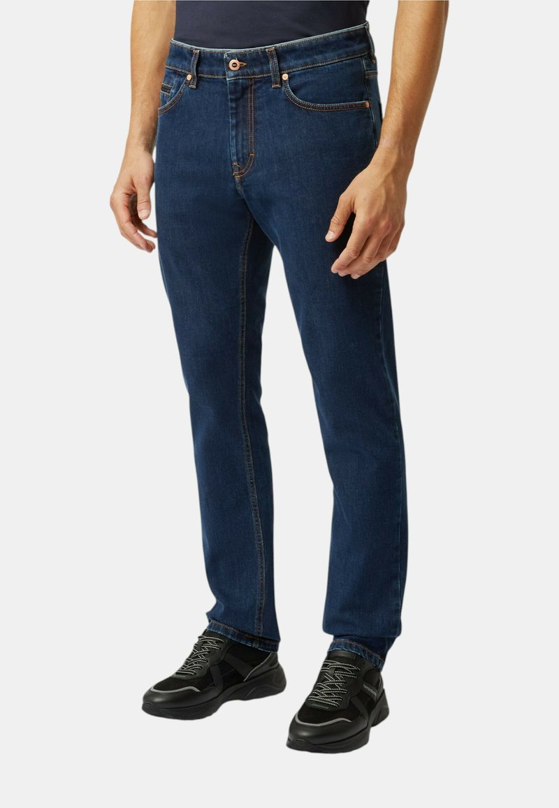 Jeans in denim blu scuro con vestibilità dritta, cuciture in vista, cinque tasche e chiusura con bottone anteriore. Indossati con scarpe sportive nere.