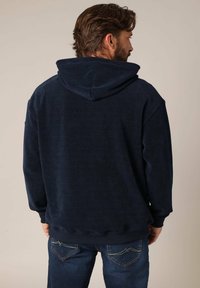 Sweatshirt à capuche bleu marine avec une texture côtelée et des manches longues. Dispose d'une capuche spacieuse et d'une taille ajustée. Porté avec un jeans bleu foncé.