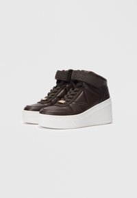 Bruine hoge sneakers met dikke witte platformzolen, vetersluiting en klittenbandsluitingen met de merknaam "GUESS" erop gedrukt.