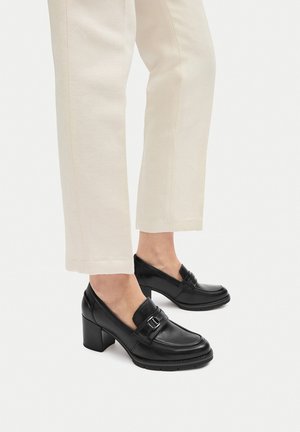 Czarne skórzane loafersy na obcasie z zaokrąglonym noskiem i ozdobnym paskiem. Noszone z kremowymi spodniami, ukazujące gładką teksturę i przemyślany krój.