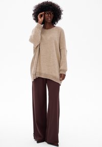 Beżowy sweter z dzianiny z oversize'owymi rękawami, w połączeniu z szerokimi brązowymi spodniami. Teksturowana tkanina, luźny krój i duże okulary w detalach.