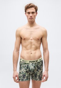 Νεαρός άνδρας με σγουρά μαλλιά που φοράει καμουφλάζ μποξεράκια Nike, μπροστά σε απλό λευκό φόντο, όρθιος και κοιτάζει μπροστά.