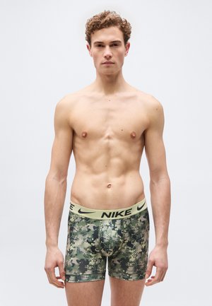 Νεαρός άνδρας με σγουρά μαλλιά που φοράει καμουφλάζ μποξεράκια Nike, μπροστά σε απλό λευκό φόντο, όρθιος και κοιτάζει μπροστά.