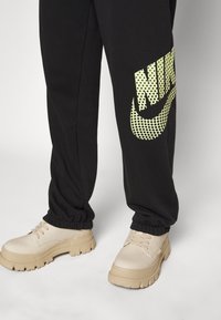 Pantalones de chándal negros con un gran logo de Nike de puntos amarillos en la pierna izquierda, combinados con zapatos beige de suela gruesa con una textura suave.