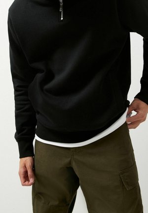 Osoba ubrana w czarną bluzę z półzamkiem na suwak, na białą koszulę i oliwkowe bojówki, poprawiająca brzeg koszuli.