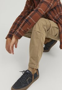 Hnědá kostkovaná flanelová košile, béžové cargo kalhoty a tmavě modré tenisky. Oblečení má strukturovanou látku a praktické kapsy.