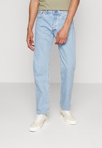 Lichtblauwe spijkerbroek met een relaxte pasvorm, voorzien van vijf zakken en contrasterende stiksels. Gecombineerd met beige sneakers met bloemaccenten.