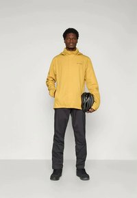 Sárga vízálló kabát kapucnival, elülső cipzárral és logóval. Fekete nadrággal és cipővel párosítva, fekete kerékpáros sisakot tart.