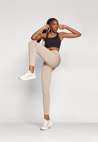 Sujetador deportivo negro, mallas beige acanaladas y zapatillas blancas. Las mallas tienen una cintura alta y una silueta ajustada. El modelo está en una pose dinámica.