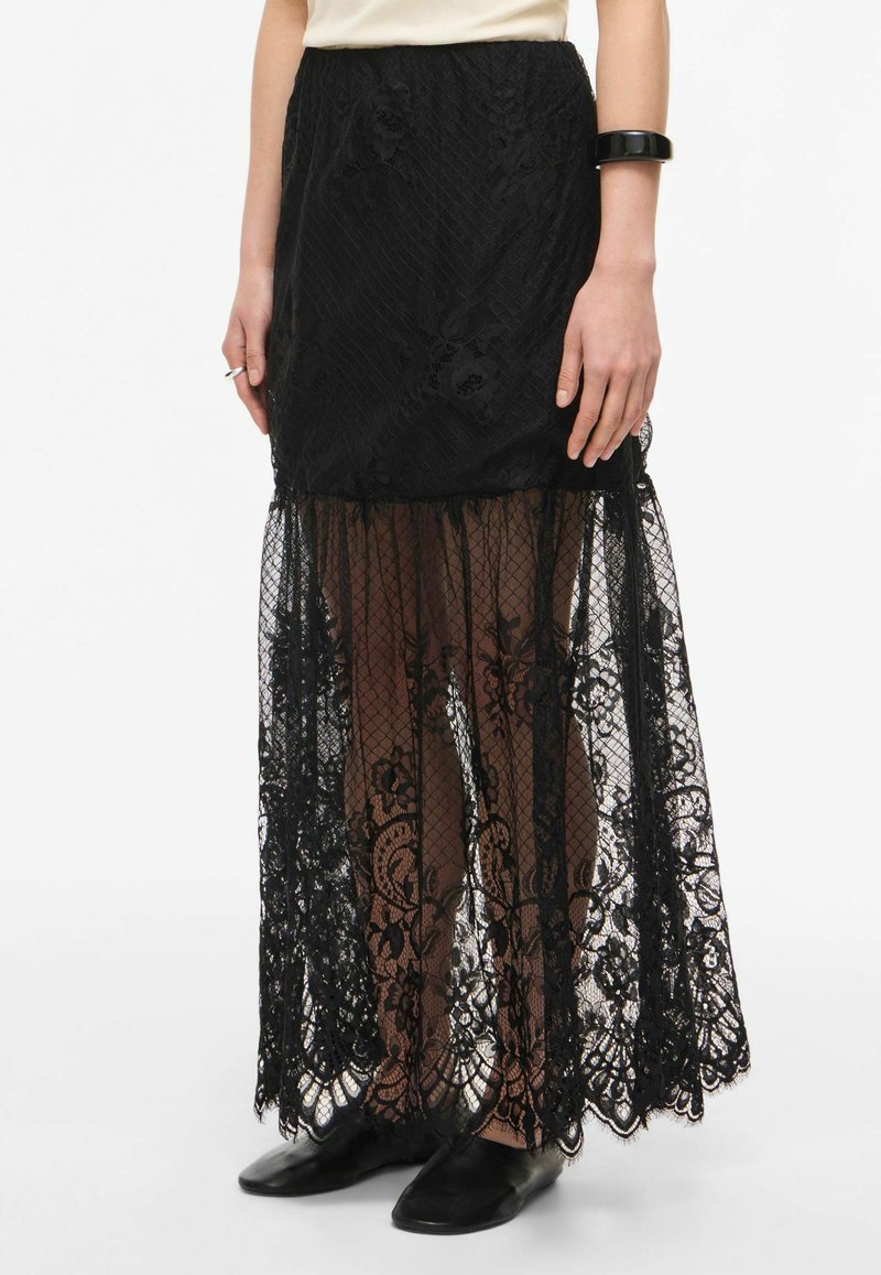 Jupe en dentelle noire avec motifs floraux et tulle transparent s'étendant sous la doublure opaque, portée avec des chaussures plates noires et un haut crème.