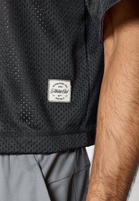 Μαύρο αθλητικό τοπ από δίχτυ με τετράγωνη ετικέτα επωνυμίας που αναγράφει «Ιδρύθηκε το 1972» και «Απλώς κάν' το». Μερικώς ορατά γκρι υφασμάτινα σορτς.