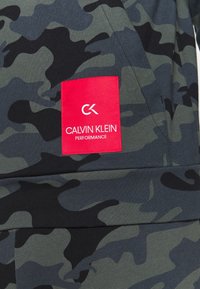 Hoodie Calvin Klein Performance présentant un motif camouflage dans des nuances de vert et de noir, avec un patch logo rouge à l'avant.