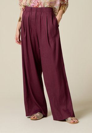 Femme portant un pantalon prune taille haute à jambes larges, associé à des sandales tressées dorées et un chemisier fleuri à manches longues avec des bracelets dorés.