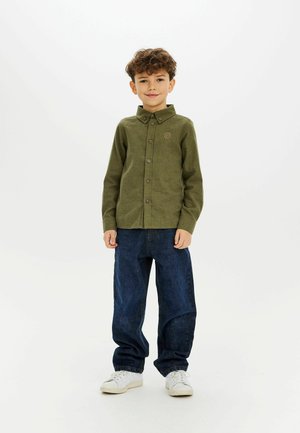 Camicia verde a maniche lunghe con una texture sottile, abbinata a jeans blu scuri dal taglio morbido e sneakers bianche.
