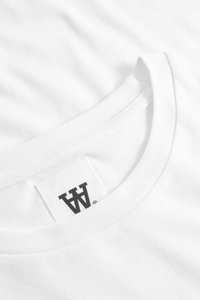 Nahaufnahme eines weißen Rundhals-T-Shirts mit einem schwarzen stilisierten "W"-Logo-Label, das im Kragen eingenäht ist.