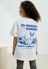 Vit bomull t-shirt med en blå design av en Mumin-karaktär i en trädgård med pumpor, texten "MUMLAND TRÄDGÅRD GLAD HÖST" nedanför.
