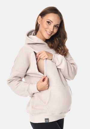 Frau in beiger Kapuzenjacke mit Reißverschluss, die einen Still-BH zeigt und lächelt, und das designfreundliche Stillkleidung präsentiert.