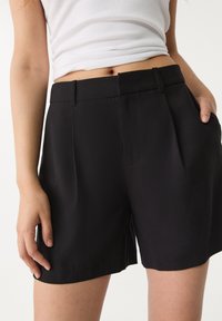 Černé šortky z hladké látky mají přizpůsobený střih s páskem v pase, předními sklady a postranními kapsami.