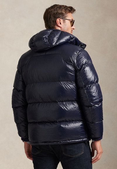 Man wearing dark navy shiny puffer jacket with hood, dark jeans, and sunglasses facing away against a plain background.