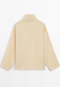 Beige top met lange mouwen, hoge kraag, losse pasvorm en zichtbare middennaad aan de achterkant, afgebeeld tegen een effen witte achtergrond.