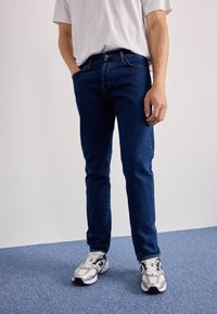 Jeans i mørkeblått denim, slim fit-design med frontlommer og kontrasterende sømmer, sammen med grå og svarte treningssko.