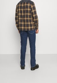 Camisa de franela a cuadros en tonos negro, marrón y amarillo, combinada con vaqueros de denim azul y calzado negro. Vista trasera mostrando detalles.