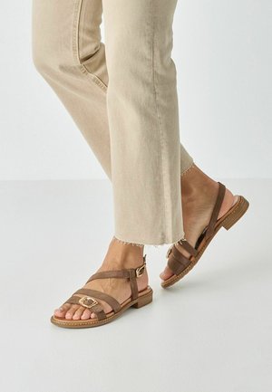 Pantalon beige raccourci avec ourlets effilochés, porté avec des sandales plates marron ornées de boucles dorées, sur un fond clair.