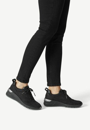 Person trägt schwarze, eng anliegende Hose und schwarze Schnürsneaker mit grauen Sohlen, steht mit einem leicht angehobenen Fuß.