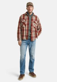 Beige karierte Flanellbluse mit roten und schwarzen Akzenten, grauer Strickpullover, blaue Jeans, braune Stiefel und eine helle Kappe.