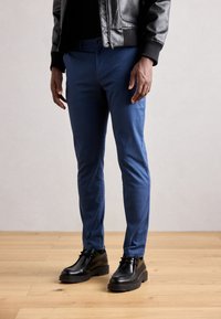Chinos azules de algodón con un corte ajustado y una textura suave, combinados con zapatos negros de cordones y una chaqueta de cuero negra.
