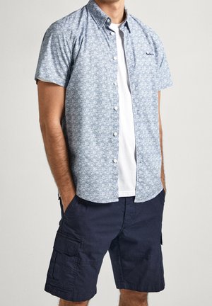 Uomo che indossa una camicia azzurra chiara con maniche corte e fantasia, sbottonata, sopra una maglietta bianca e pantaloncini cargo blu scuro, con le mani nelle tasche.