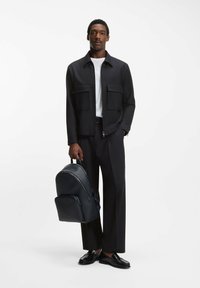 Svart lättviktsjacka med två framfickor, vit t-shirt, svarta byxor och en texturerad mörk ryggsäck. Svarta skor fulländar looken.