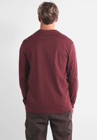 Chemise à manches longues couleur bordeaux en coton, présentant un design simple avec un col arrondi et un petit logo sur l'épaule gauche.