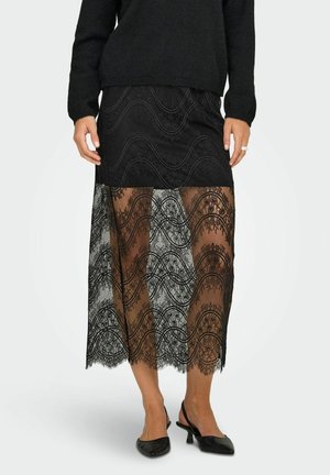 Jupe maxi en dentelle noire avec un motif en vagues, couche inférieure semi-transparente et partie supérieure noire unie. Associée à des chaussures à talons bas noires.