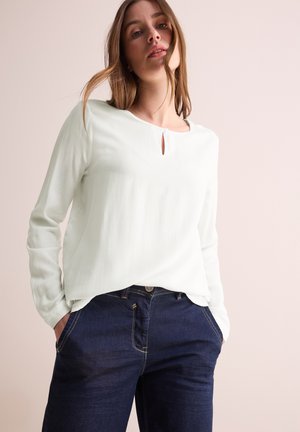 Blouse à manches longues, vert clair, en tissu texturé avec un détail en forme de trou de serrure, associée à un jean en denim bleu foncé.