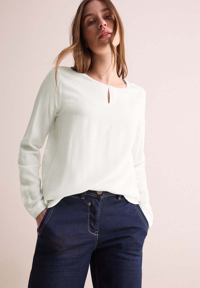 Blusa a maniche lunghe di colore verde chiaro con tessuto testurizzato e dettaglio a forma di chiave sul collo, abbinata a jeans di denim blu scuro.