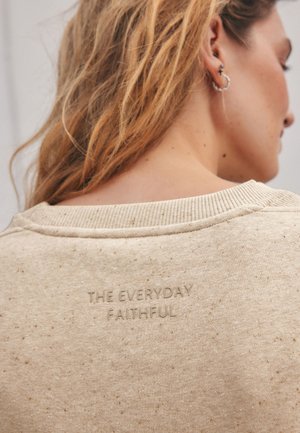 Femme aux longs cheveux blonds portant un sweat-shirt beige moucheté avec l’inscription en relief « THE EVERYDAY FAITHFUL » dans le dos, près du col.