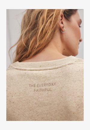 Femme aux longs cheveux blonds portant un sweat-shirt beige moucheté avec l’inscription en relief « THE EVERYDAY FAITHFUL » dans le dos, près du col.