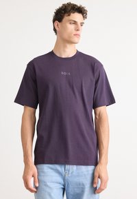 T-shirt en coton violet à manches courtes et col à ras du cou. Doté d'un logo central "BOSS" dans une teinte plus claire. Porté avec un jean léger bleu.