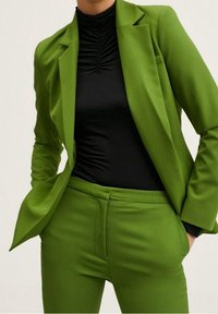 Blazer verde ajustado y pantalones de cintura alta a juego sobre una blusa negra texturizada, con hombros estructurados y corte ajustado.