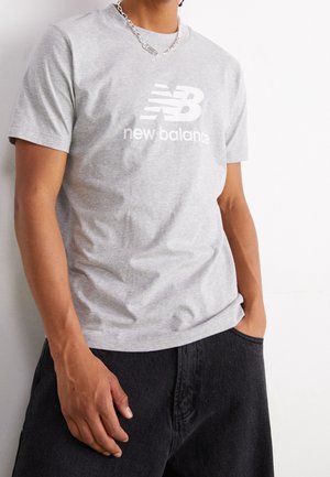 Person iført en lysegrå New Balance t-shirt og mørke bukser, stående foran en ensfarvet hvid baggrund med den ene hånd i lommen.