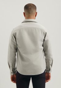 Een man in een lichtgrijze lange mouw knopshirt, gezien vanuit de rug, staand tegen een effen lichte achtergrond.