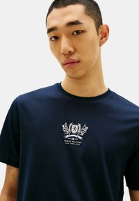 T-shirt di cotone blu navy con collo a giro, presenta un logo ricamato di Tommy Hilfiger in bianco e grigio sul petto. Texture liscia.